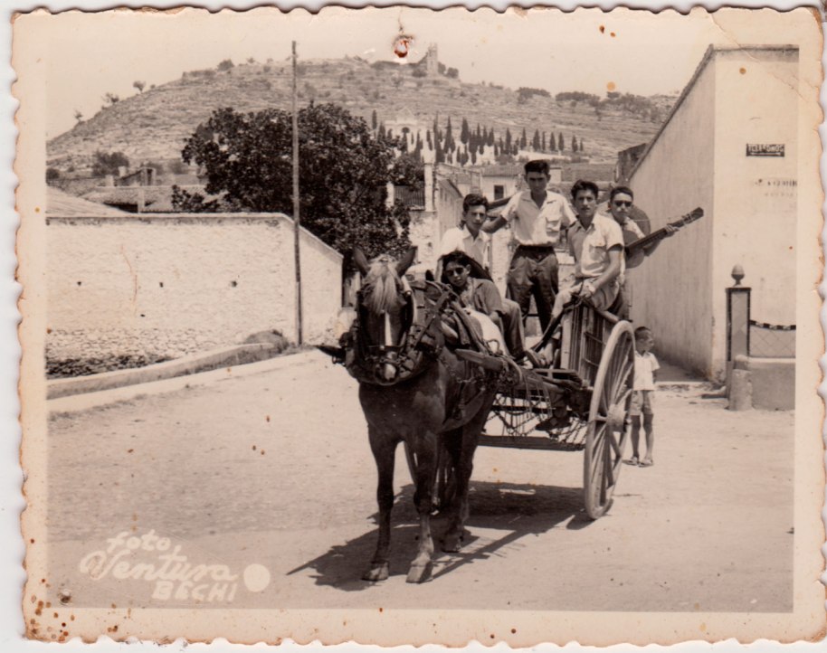 A cavall d'un carro, a migdia, amb sombreros mexicans i guitarres, possiblement anant de paella a l'Ermita. Eixen del poble pel carrer Sant Ramon. A la dreta es veu l'abeurador molt adornat que després es llevaria per posar-lo a l'altre costat de carrer (i on Roia va trencar la Reva). Els joves són, d'esquerra a dreta, Escaleta, Juanito Corina, Vicente Pla, un desconegut, i davant de tots Joaquín de Músic.